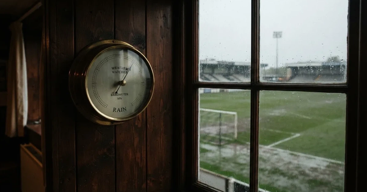 Ein Wetterbarometer hängt an einer Holzwand neben einem Fenster, draussen liegt ein nasser Fussballplatz im Regen.