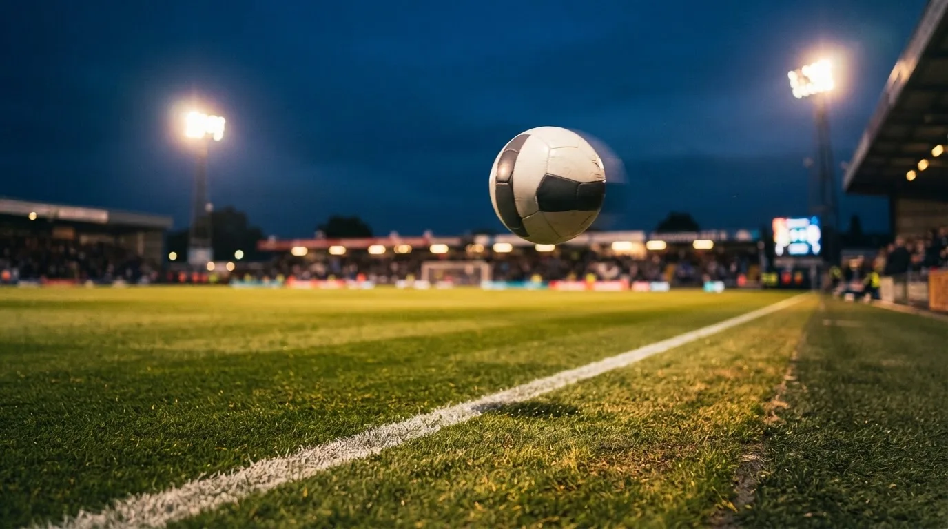 Ein Fussball fliegt in der Abenddämmerung über einen beleuchteten Rasen, aufgenommen von der Seitenlinie aus.