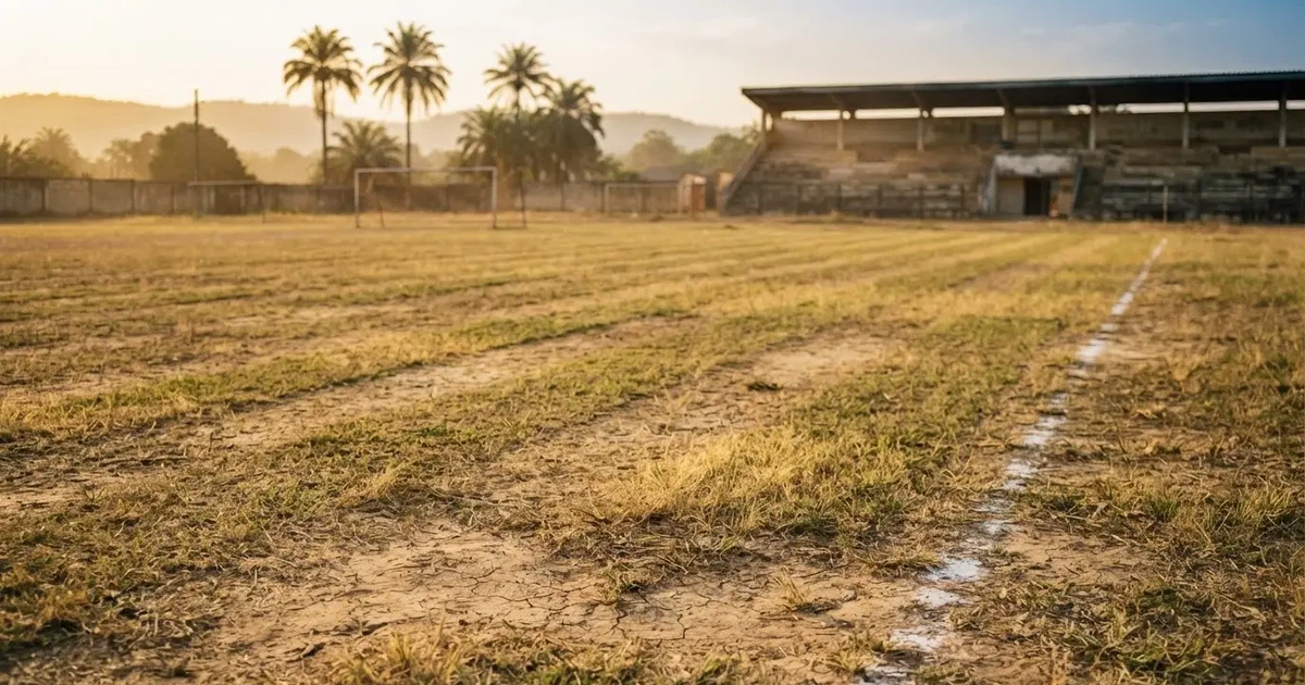 Ein ausgetrockneter Fussballplatz mit Rasenstreifen im warmen Nachmittagslicht, im Hintergrund Palmen und ein flaches Stadiondach.