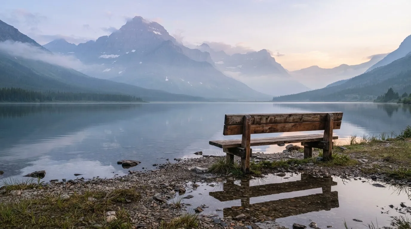 Eine Holzbank an einem Seeufer mit Blick auf eine Bergkette im Morgendunst, das Wasser ist spiegelglatt.