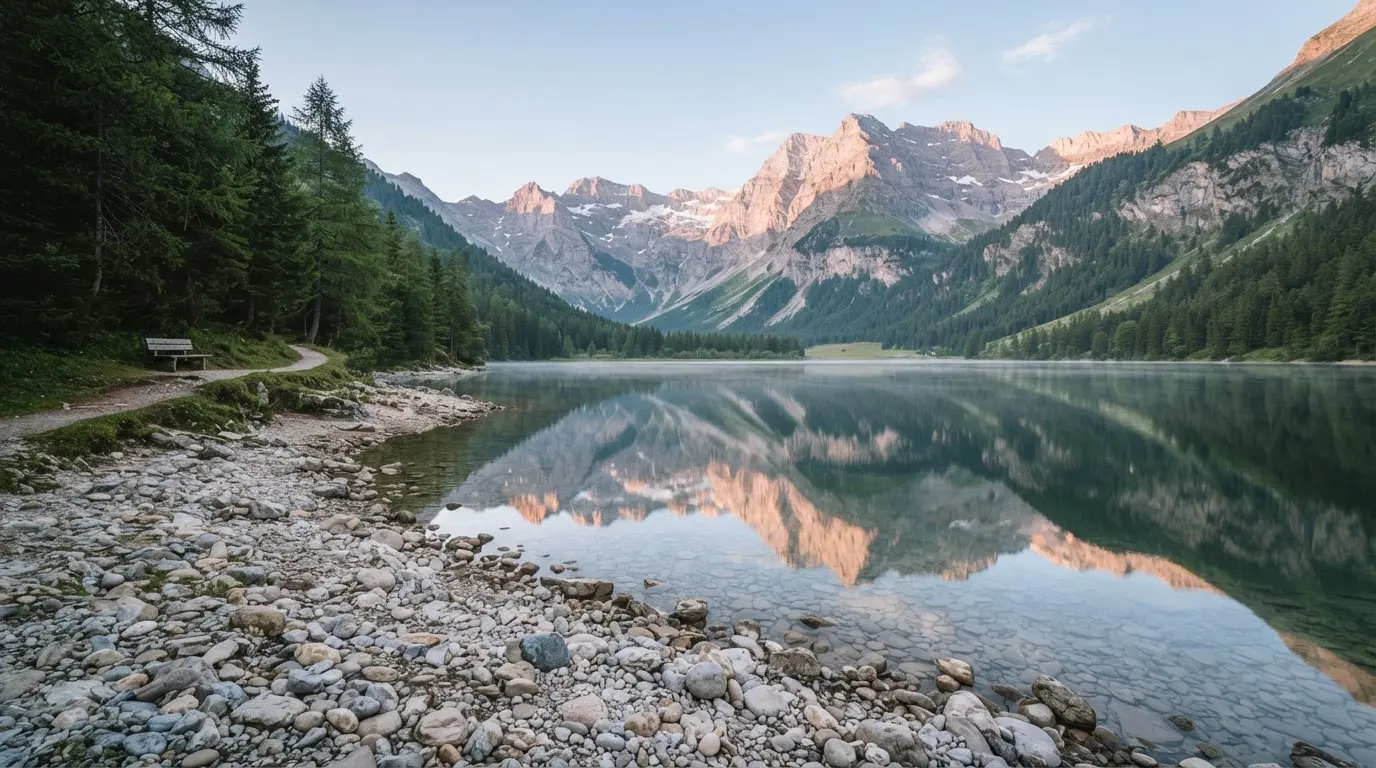 Ein Bergsee mit ruhigem Wasser und Gipfeln im Morgenlicht, am Ufer liegen glatte Steine.