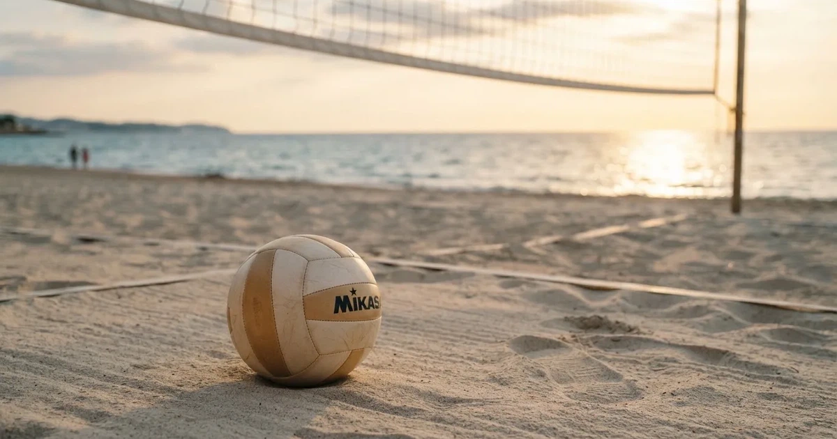 Ein Volleyball liegt auf feinem hellem Sand, ein gespanntes Netz verschwimmt dahinter, das Licht kommt von einem weiten Meer.
