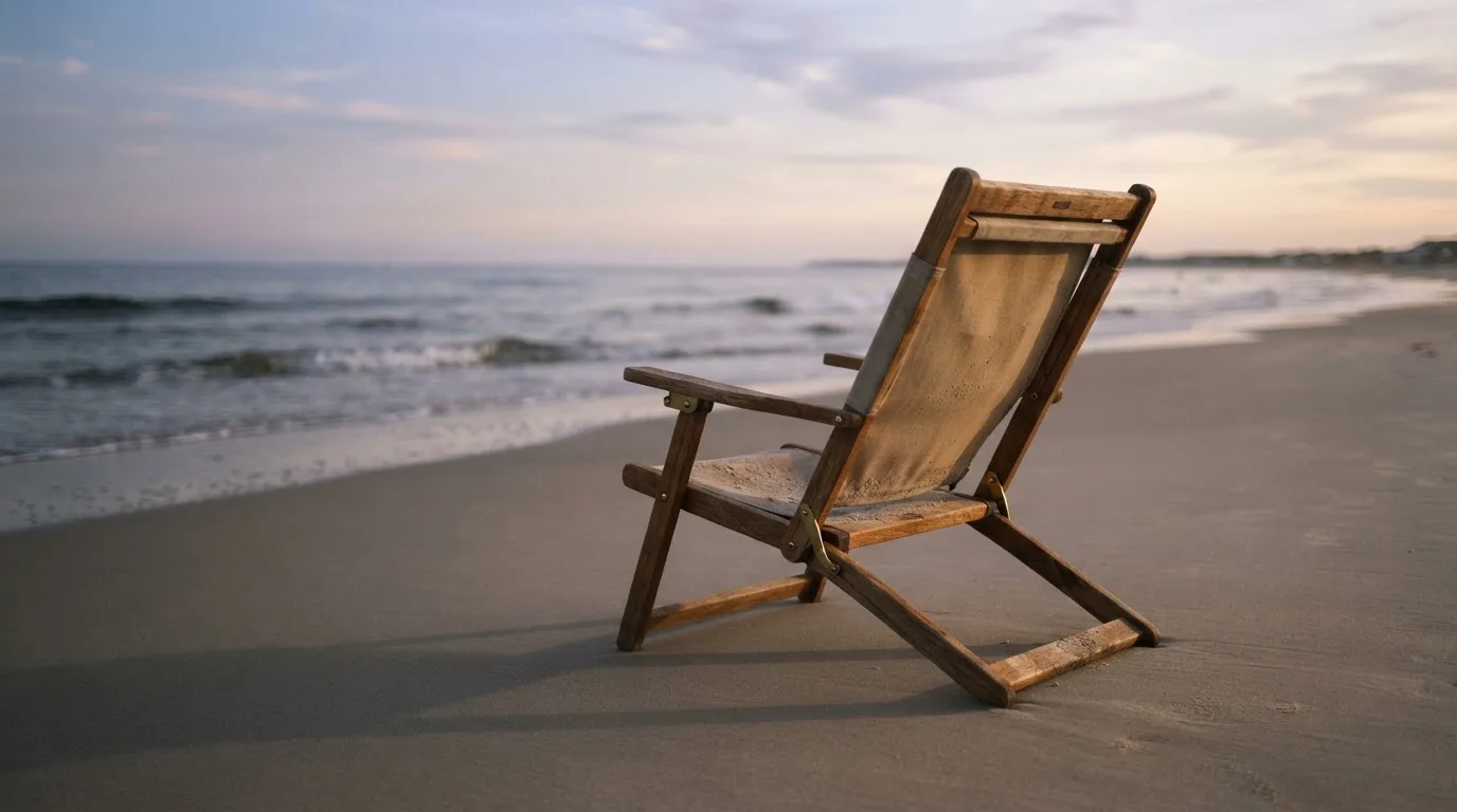 Ein leerer Liegestuhl aus Holz steht am Rand eines Sandstrands, Abendlicht und ruhige Wellen im Hintergrund.