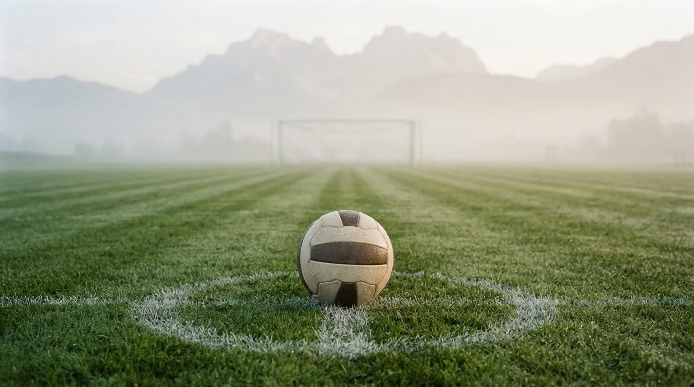 Ein einzelner Fussball auf dem Mittelpunkt eines leeren Rasens im Morgennebel, hinter dem Tor ragen Berge auf