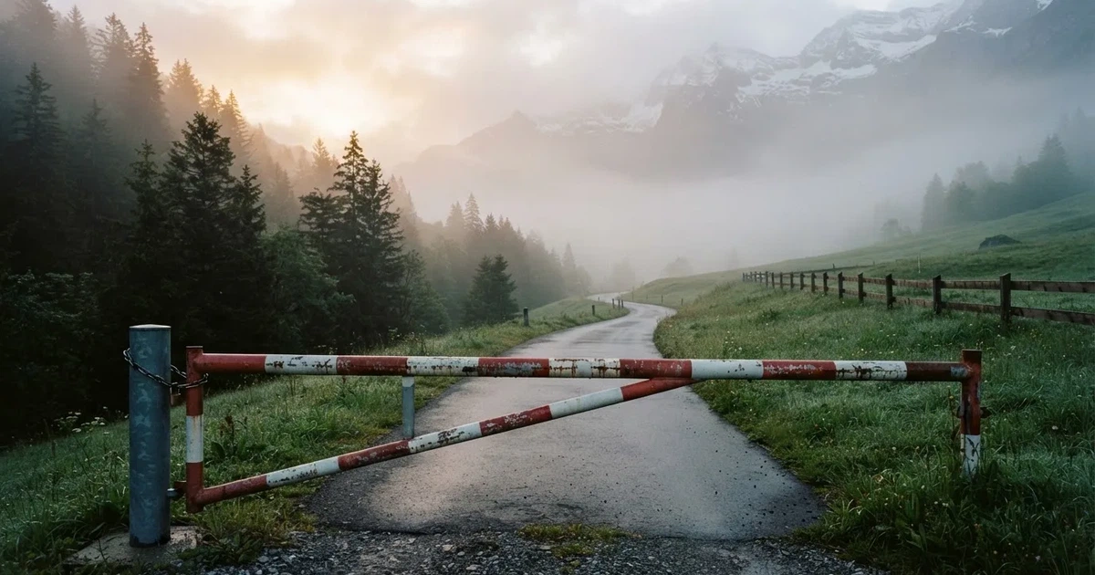 Eine geschlossene Schranke steht an einer ruhigen Bergstrasse im Morgennebel, dahinter Tannen und eine nasse Wiese.